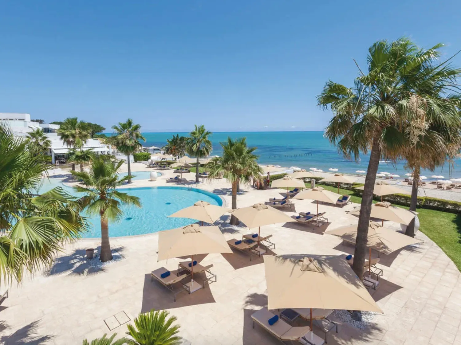 Bord de piscine d’un complexe hôtelier en front de mer, avec palmiers, transats, parasols beiges et plage de sable.